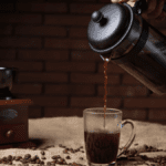 Prensa francesa cheia de café, despejando café na xícara de vidro sobre a pesa. Ao fundo, um moedor de grãos de café manual de madeira.