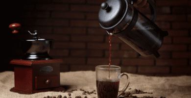 Prensa francesa cheia de café, despejando café na xícara de vidro sobre a pesa. Ao fundo, um moedor de grãos de café manual de madeira.