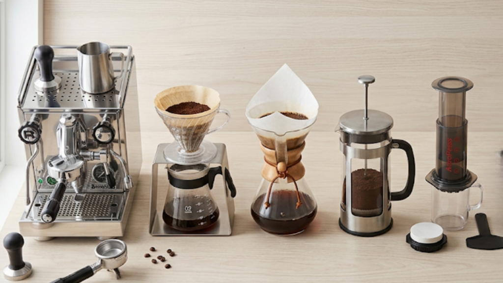 Flat lay aéreo sobre bancada de madeira clara exibindo cinco equipamentos de preparo lado a lado: máquina de espresso, V60 com suporte, Chemex, prensa francesa, Aeropress. Composição equilibrada e harmoniosa, luz natural difusa vinda da esquerda.