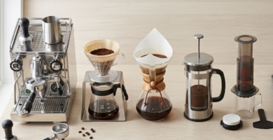 Flat lay aéreo sobre bancada de madeira clara exibindo cinco equipamentos de preparo lado a lado: máquina de espresso, V60 com suporte, Chemex, prensa francesa, Aeropress. Composição equilibrada e harmoniosa, luz natural difusa vinda da esquerda.