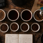 Mesa com setup completo de cupping de café em casa: tigelas de degustação, colher, balança e roda de sabores dispostos para avaliação sensorial