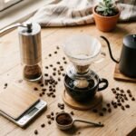 Bancada de café em casa com moedor manual, balança digital, chaleira de bico fino, coador de vidro e grãos espalhados