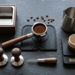 Tamper, porta-filtro, knock box, pitcher e balança organizados sobre bancada escura