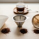 Três tipos de filtro de café lado a lado sobre madeira clara: papel, inox e pano, com café moído ao lado