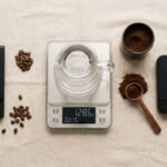 Três balanças digitais de café alinhadas sobre superfície clara com jarra de vidro e grãos