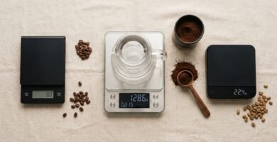 Três balanças digitais de café alinhadas sobre superfície clara com jarra de vidro e grãos