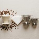 Coadores cônicos, filtros de papel e coadores de inox alinhados sobre superfície clara com jarra de vidro