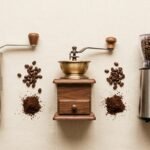 Moedores de café manuais e elétricos alinhados sobre superfície clara com grãos e café moído