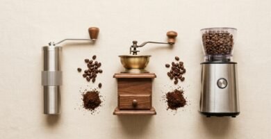 Moedores de café manuais e elétricos alinhados sobre superfície clara com grãos e café moído